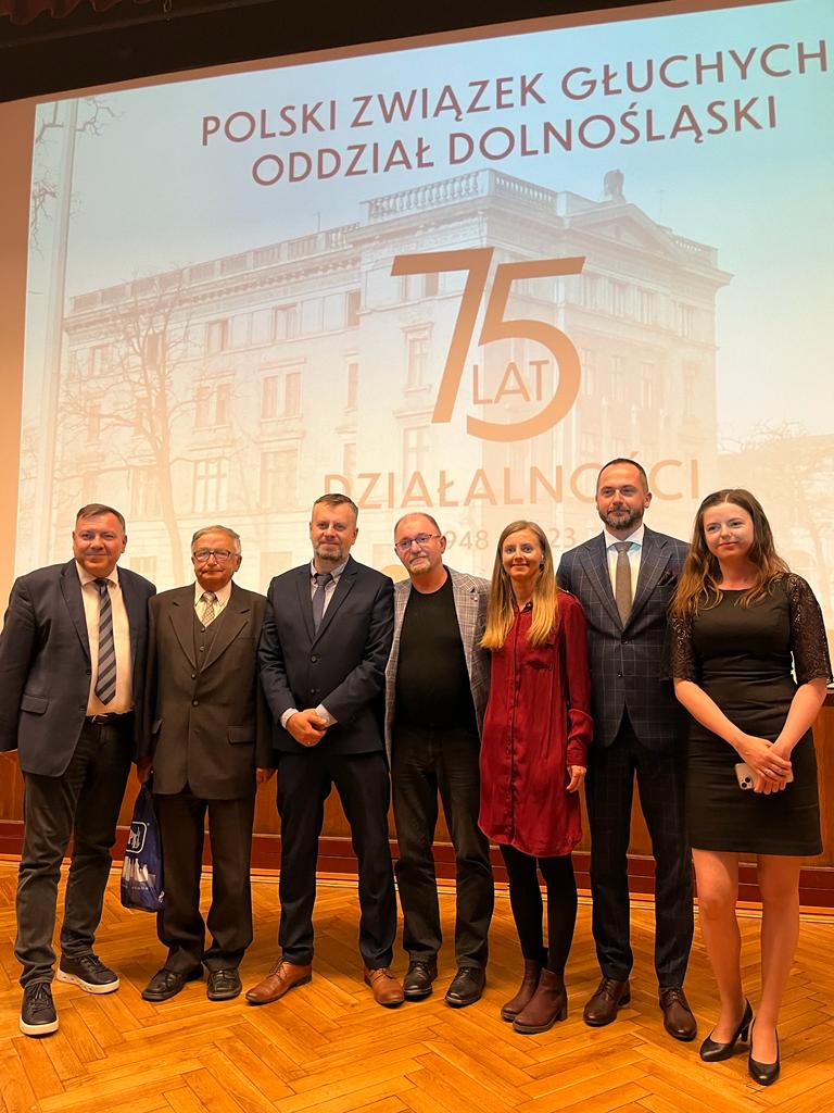 Zdjęcie przedstawia 7 osób stojących przed ekranem z napisem 75-lecie Polskiego Związku Głuchych Oddział Dolnośląski we Wrocławiu 